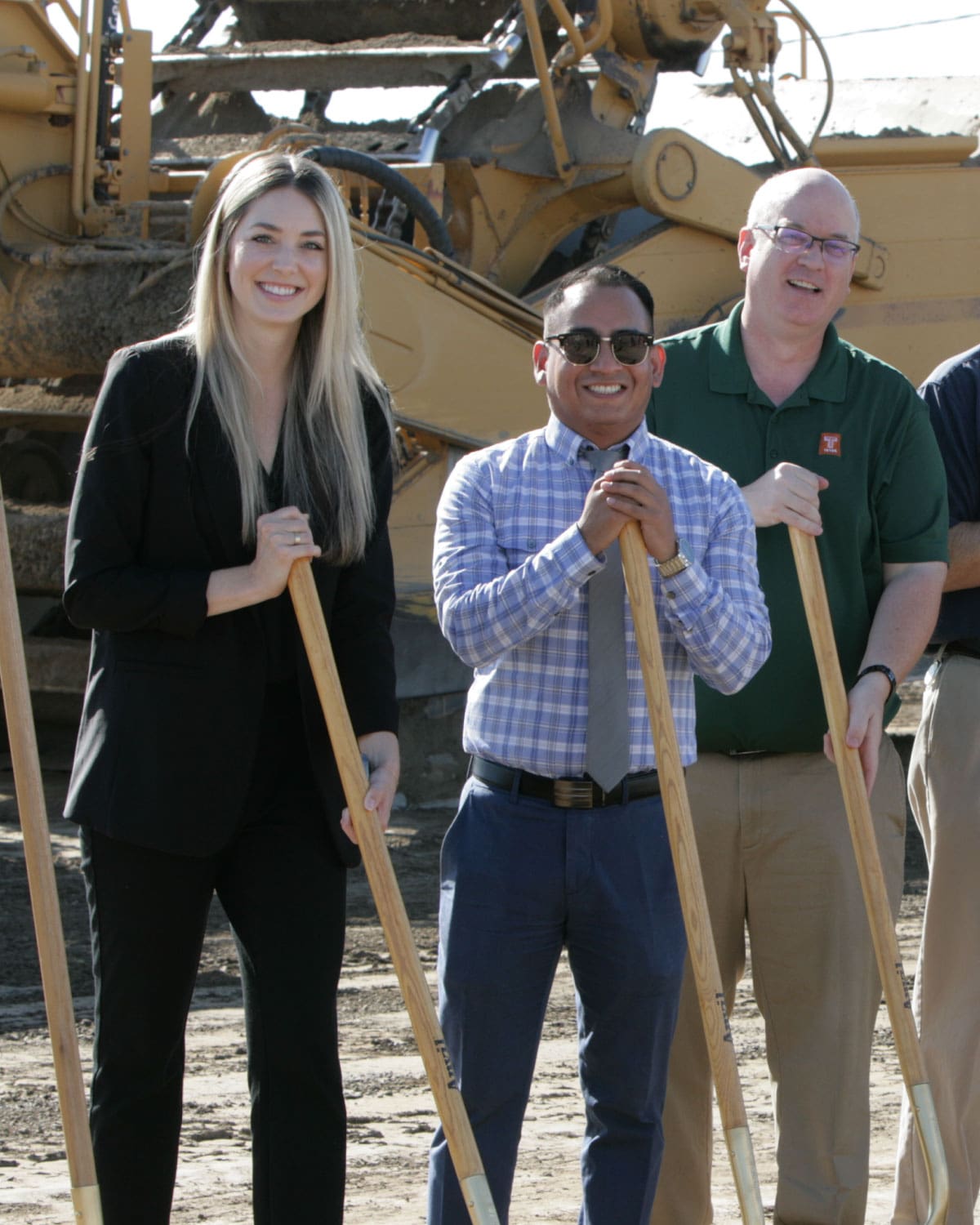 Three Teter employees holding shovels, smiling at a construction groundbreaking site with heavy machinery in the background.