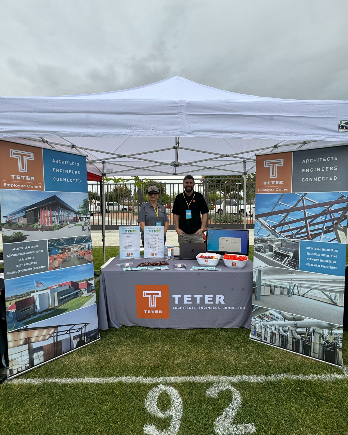 Two Teter employees standing behind a branded booth under a white tent at an outdoor event, showcasing architectural and engineering services.