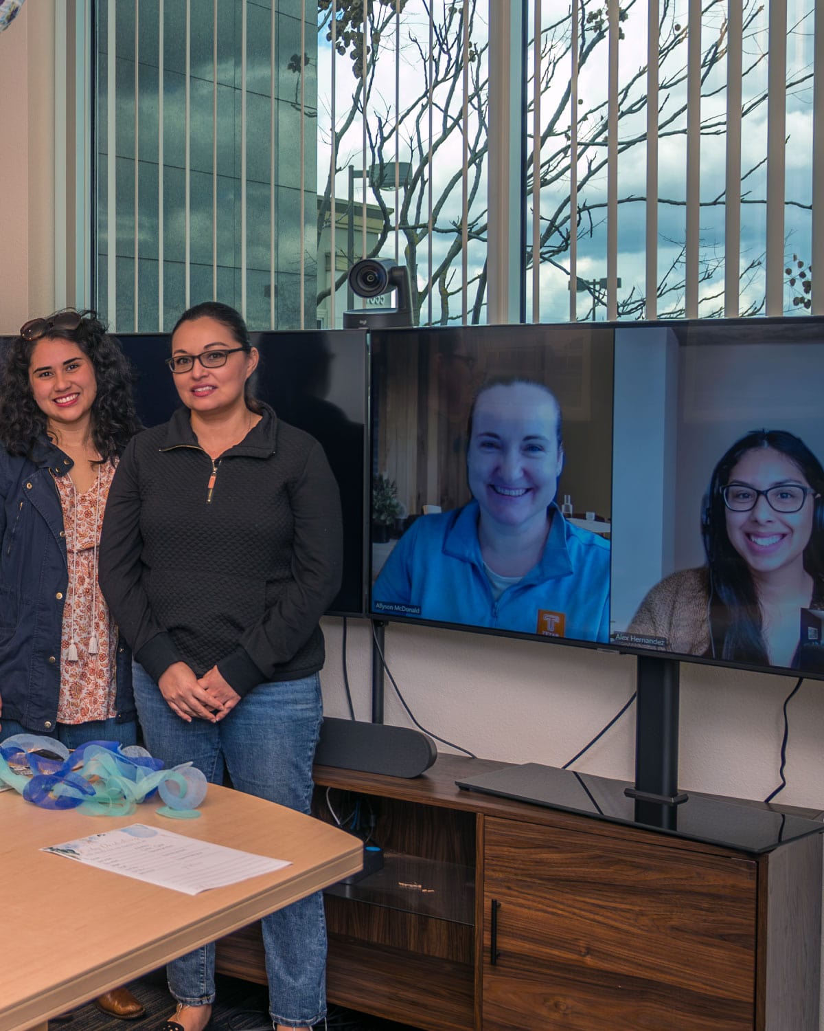 Two Teter employees standing beside a video conference screen showing remote teammates, with an office window and cityscape visible behind them.