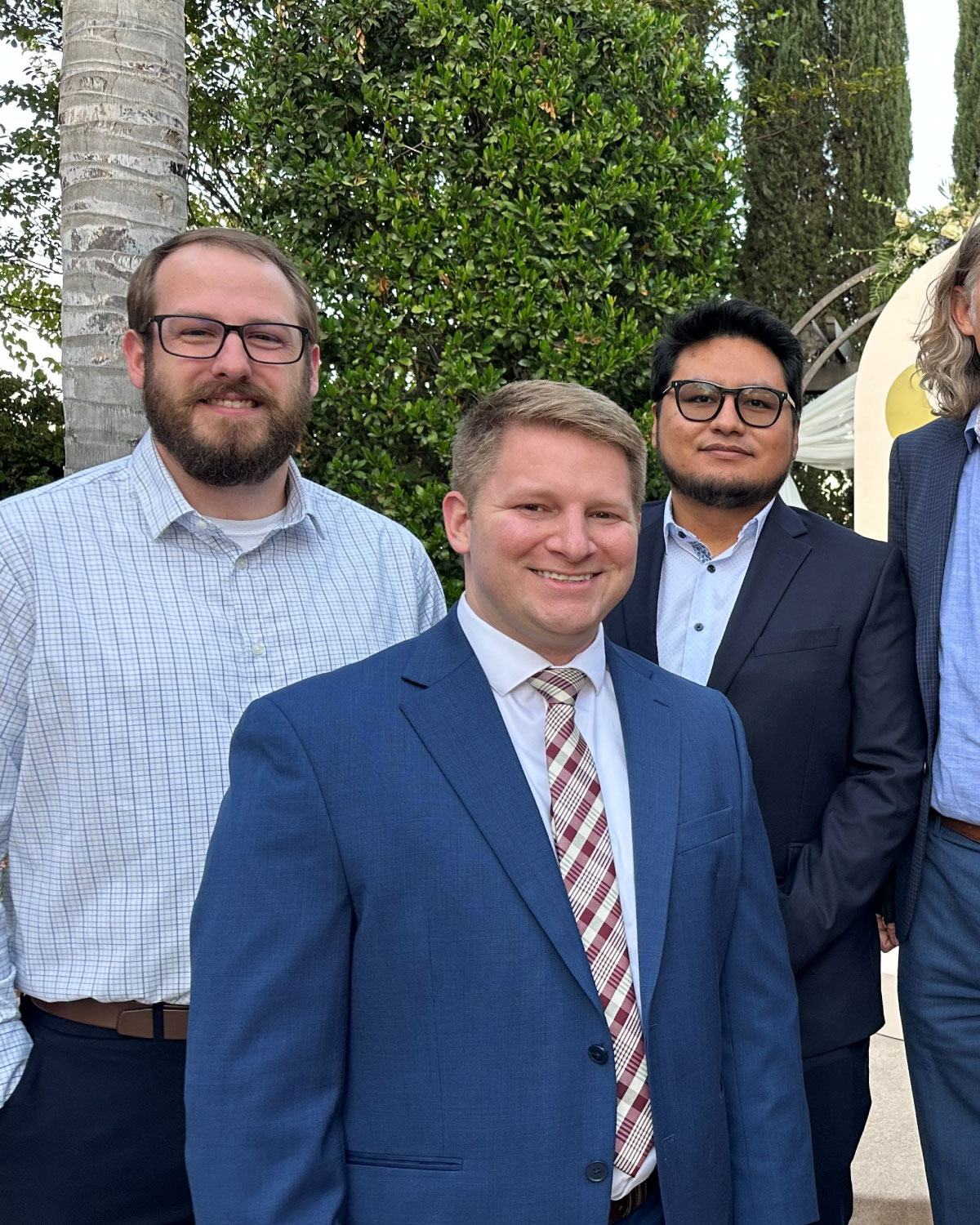 Three men in dress shirts and blazers standing outdoors, smiling against a background of leafy shrubs and a tall palm tree.