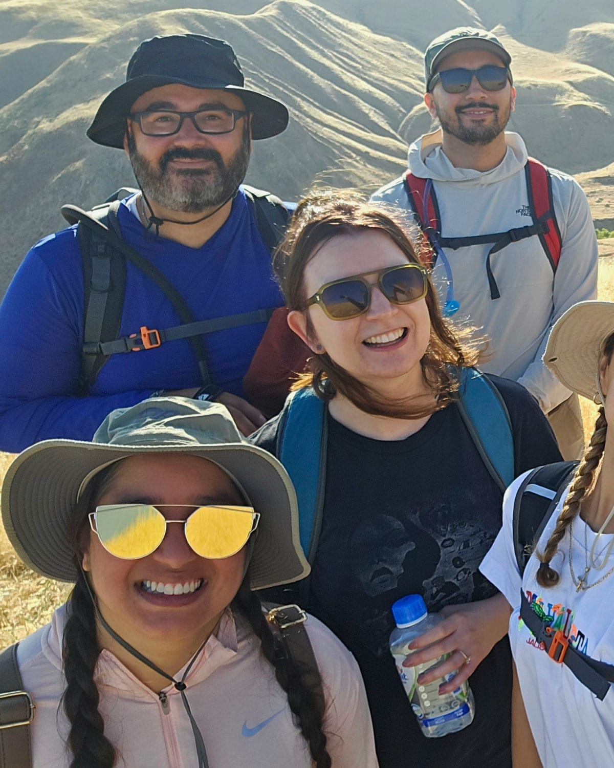 A group of Teter employees wearing outdoor gear, smiling and standing together on a hiking trail with rolling hills in the background.