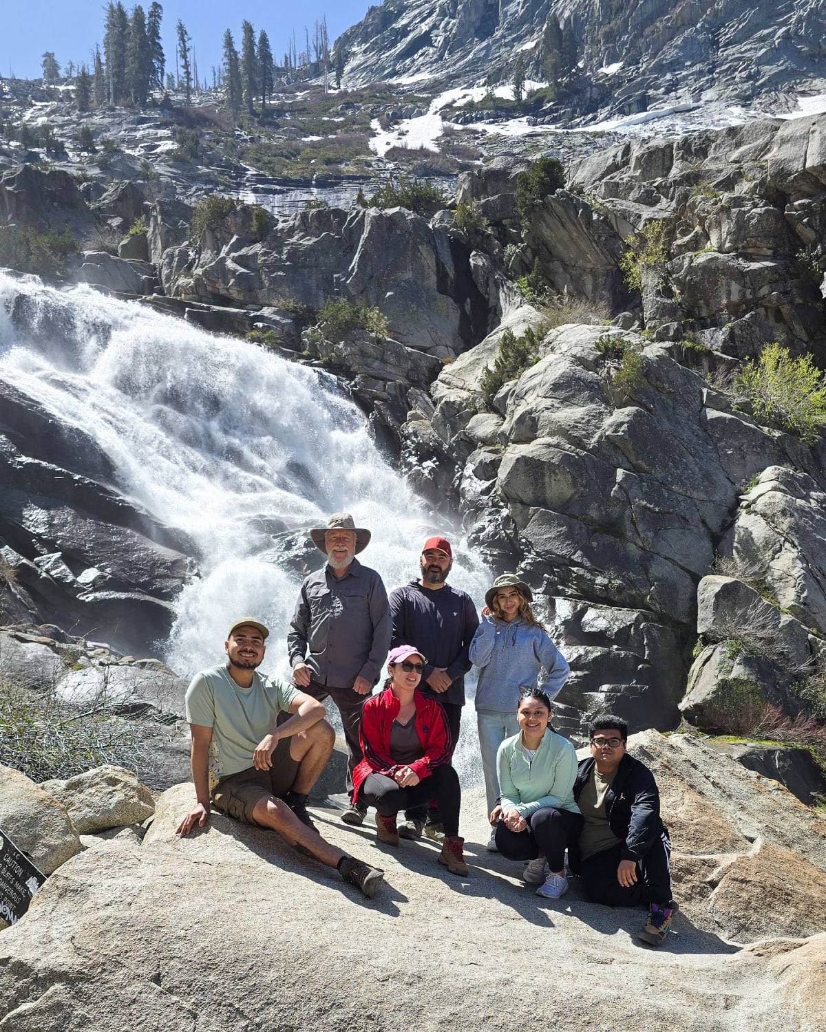 A group of Teter employees posing on a large rock in front of a rushing waterfall and rugged, snow-dusted mountain terrain.