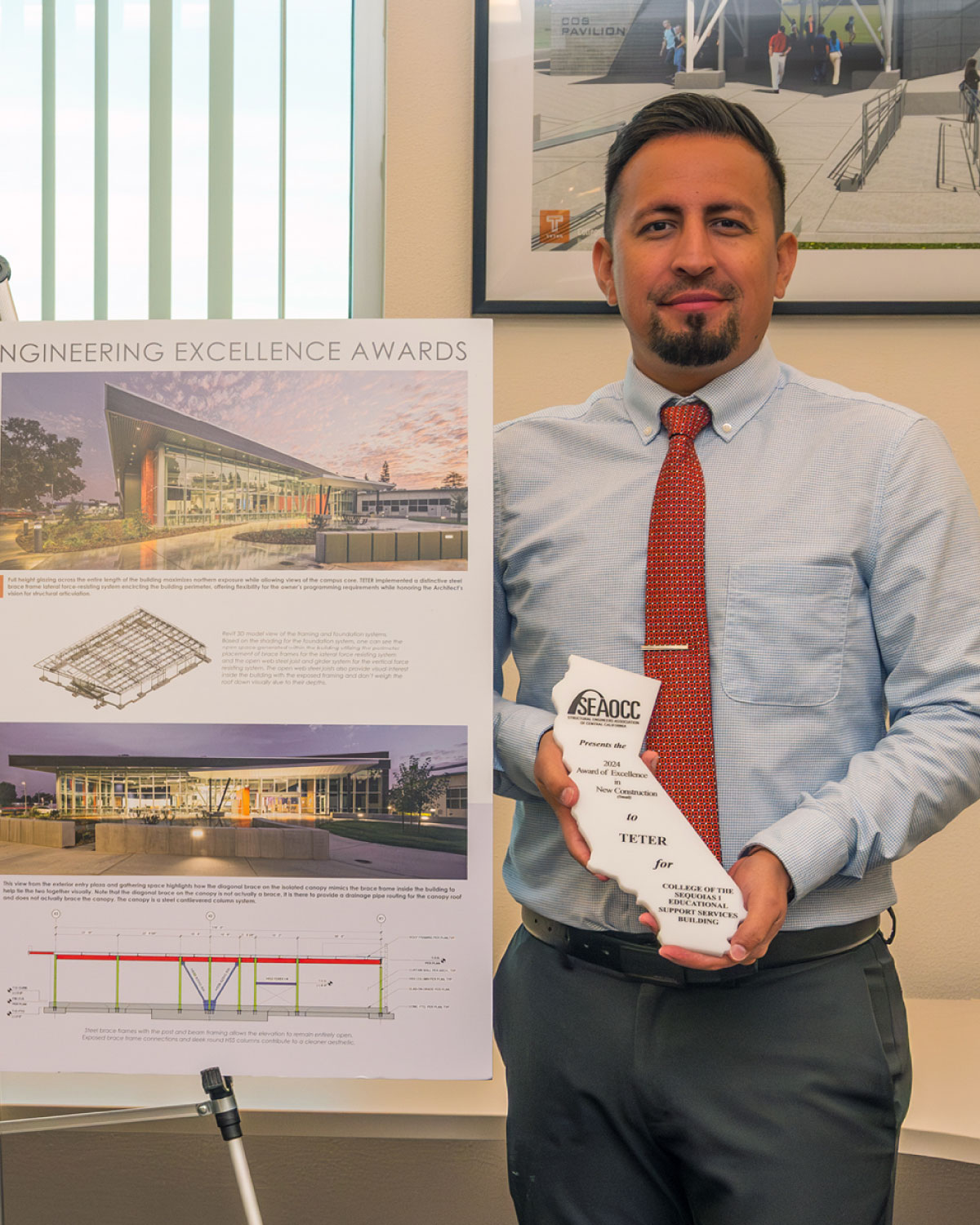 A Teter employee in a dress shirt and red tie holding an engineering excellence award and standing next to a project display board.